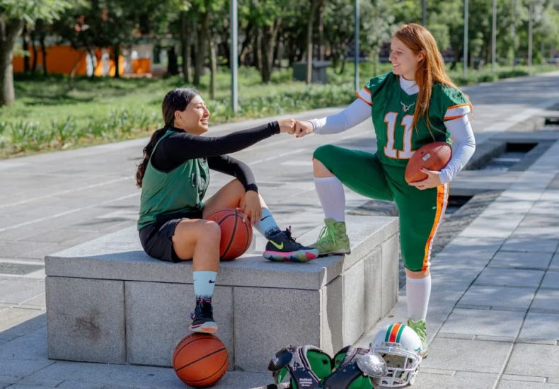 Two female athletes share a fist bump while sitting outdoors, holding sports equipment in Mexico City.